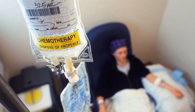 A chemotherapy IV bag is in focus, labelled CHEMOTHERAPY DISPOSE OF PROPERLY, with a person sitting in a chair receiving treatment in the blurred background.