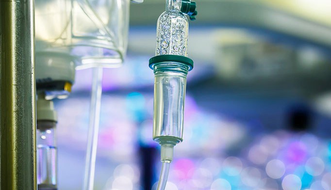 Close-up of an intravenous (IV) drip chamber with clear fluid, used for medical infusion, against a blurred background with blue and purple lights.