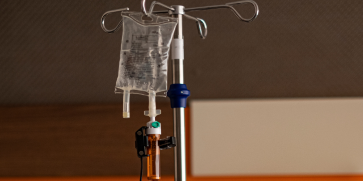 A close-up of a drip bag hanging on a metal stand indoors, with tubing attached and fluid visible inside the bag.