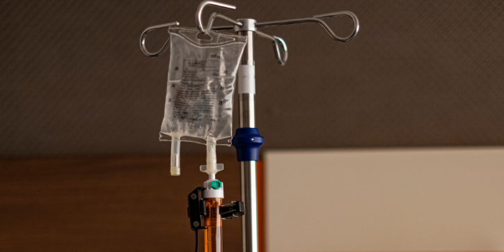 An intravenous (IV) fluid bag hangs from a metal IV pole, with attached tubing and connectors, in a medical setting under indoor lighting.