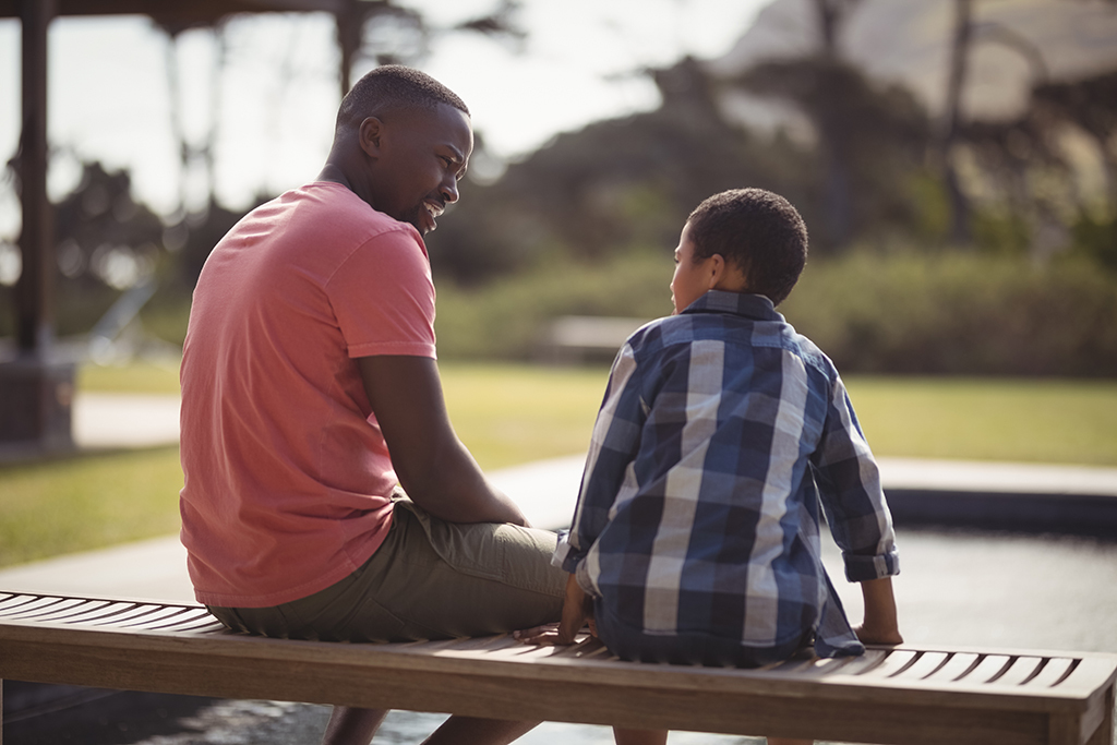 A man and a young boy sit on a wooden bench by a swimming pool, facing each other and talking outdoors on a sunny day, with greenery and hills in the background.