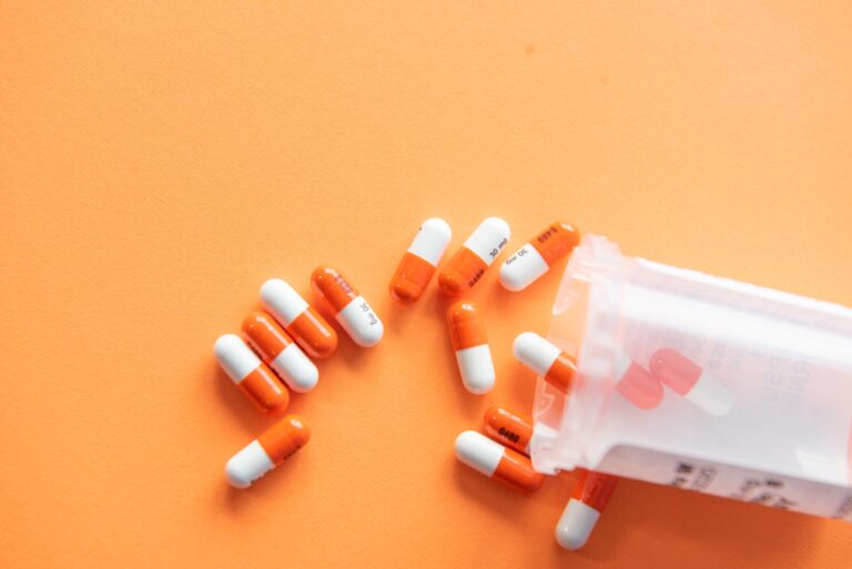 Several orange and white capsules spill out from a translucent prescription pill bag onto a solid orange background.