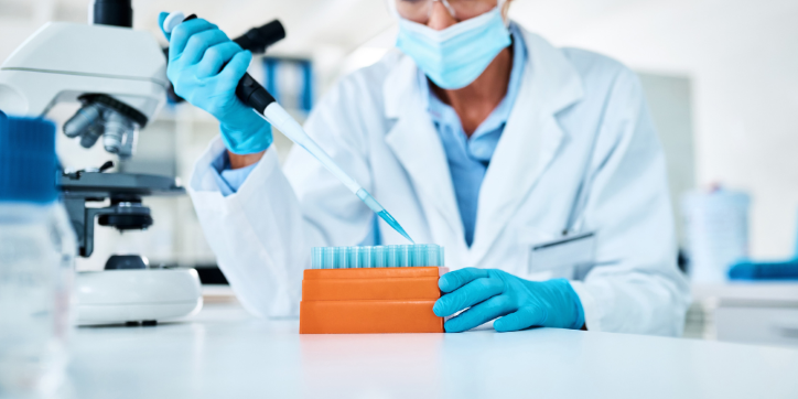 A scientist in a lab coat and mask uses a pipette to fill test tubes. A microscope and other laboratory equipment are visible on the white worktop.