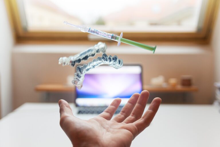 A hand throws clear dental aligners and a dental syringe into the air, with a blurred laptop and window in the background.