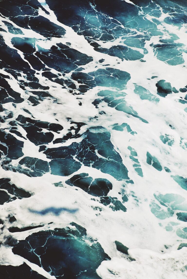 Aerial view of ocean water with swirling white sea foam and deep blue-green waves, capturing the dynamic and textured surface of the sea.
