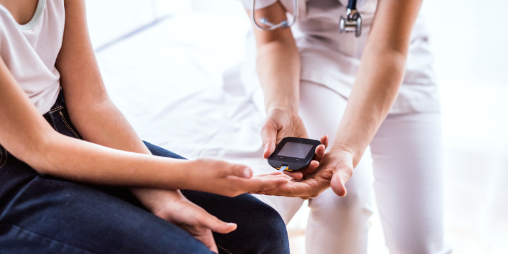 A healthcare professional holds a blood glucose metre while assisting a patient in checking their blood sugar level. The patient extends their hand for testing in a clinical setting.