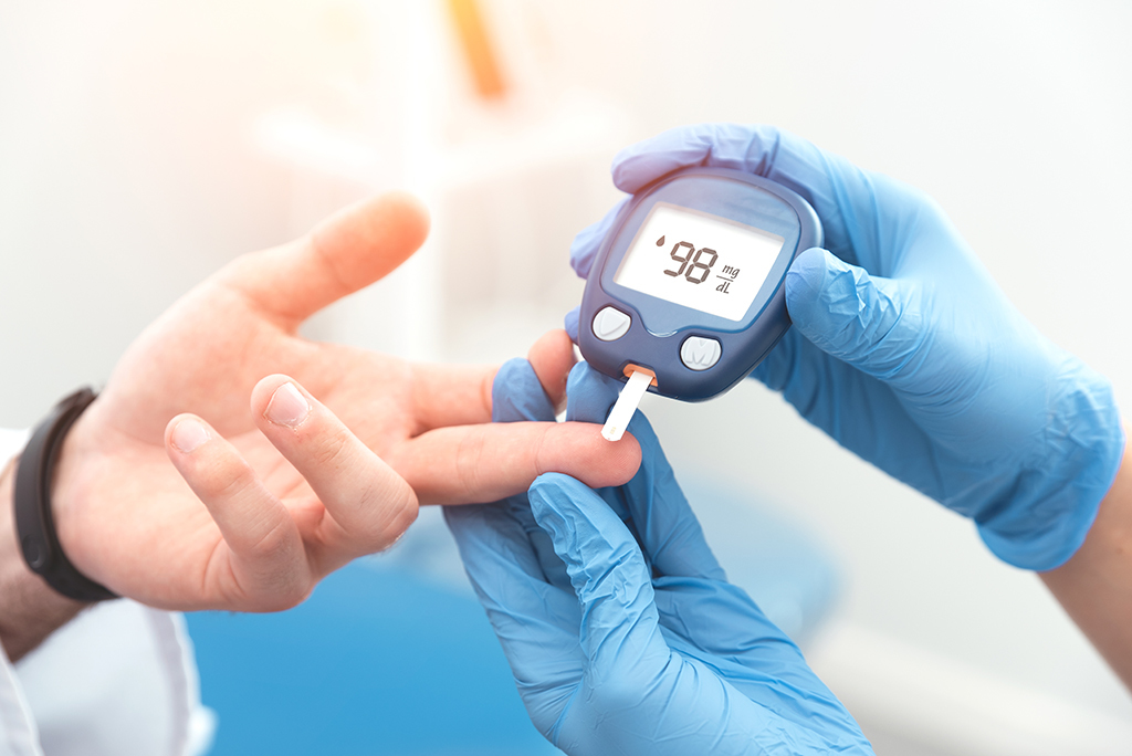 A gloved hand uses a blood glucose meter to test a person's blood sugar level by pricking their finger. The metre displays a reading of 98.
