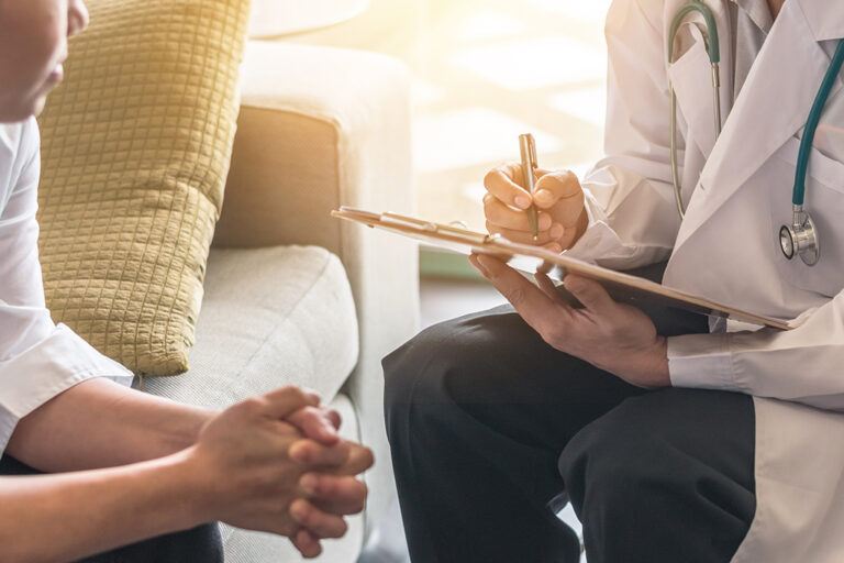 A doctor holding a clipboard and pen speaks with a seated patient. The patient's hands are clasped, and only part of their body is visible. The setting appears to be a comfortable, informal room with a sofa and cushions.