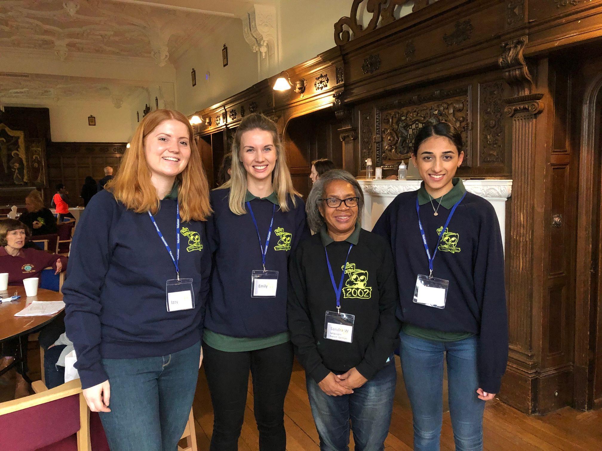Four women wearing navy sweatshirts and name badges stand together, smiling, in a wood-panelled room with ornate decor. Other people are seated at tables in the background.