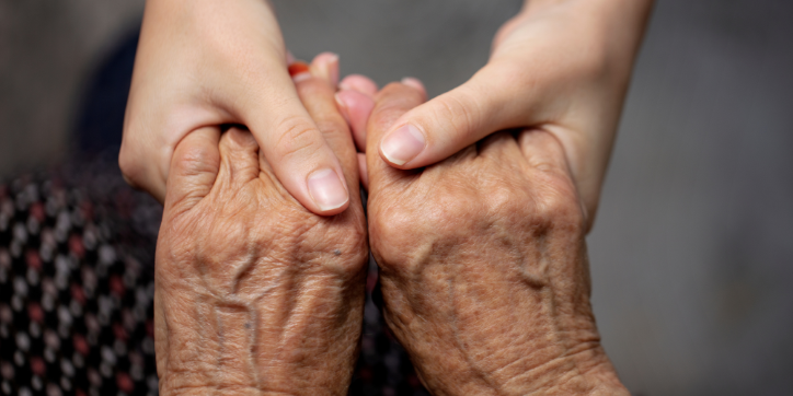 A younger person gently holds the hands of an older adult, conveying care and support. The older hands are wrinkled whilst the younger hands are smooth.