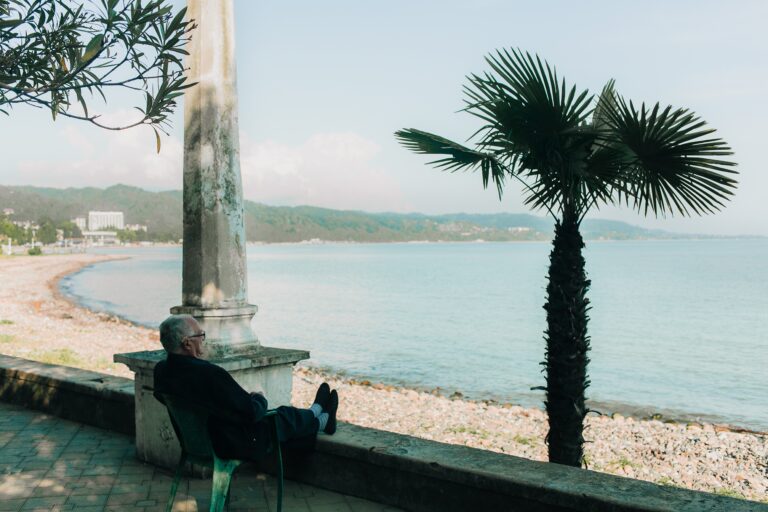 An older man sits and relaxes by a stone pillar, facing a calm beach with pebbles and a palm tree, overlooking the sea and distant green hills on a clear day.