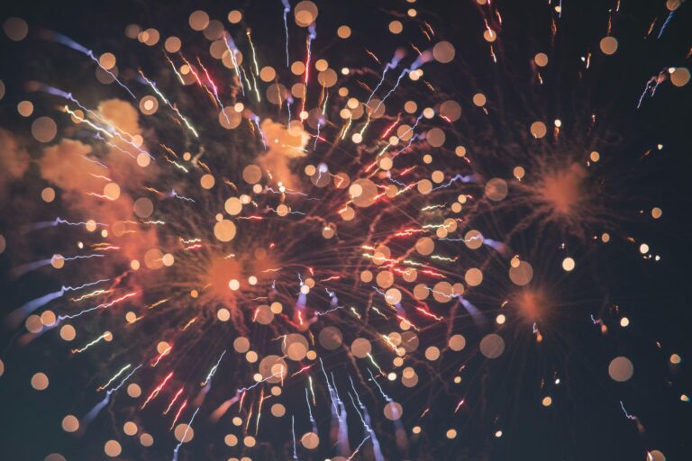 Colourful fireworks burst against a dark night sky, with bright streaks, sparks, and circular light bokeh effects scattered throughout the image.