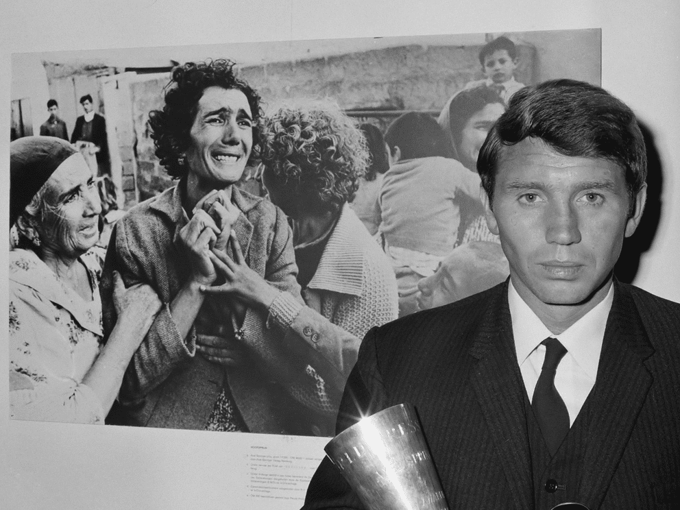 A man in a suit holds a trophy, standing in front of a black-and-white photo of distressed women and children reacting emotionally.
