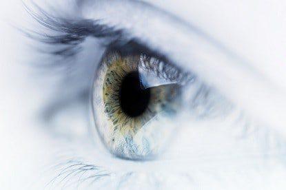 Close-up of a human eye with blue-green iris and long eyelashes, set against a bright, softly blurred white background.