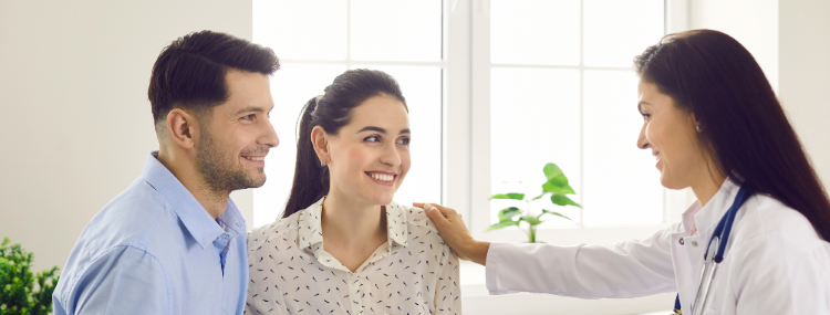 A doctor in a white coat smiles and talks with a smiling couple seated in her surgery. The doctor gently rests her hand on the woman's shoulder. A window and a potted plant are visible in the background.