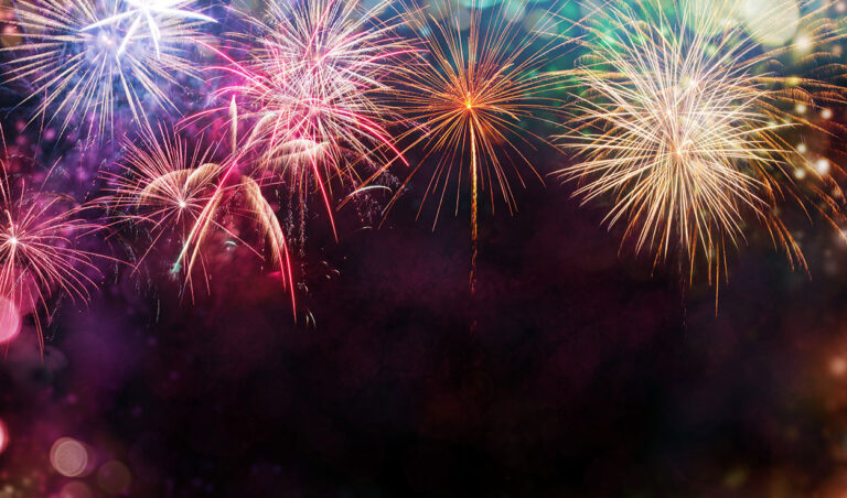 Colourful fireworks burst in the night sky, with vibrant red, yellow, blue, and orange streaks lighting up the dark background, creating a festive and celebratory atmosphere.