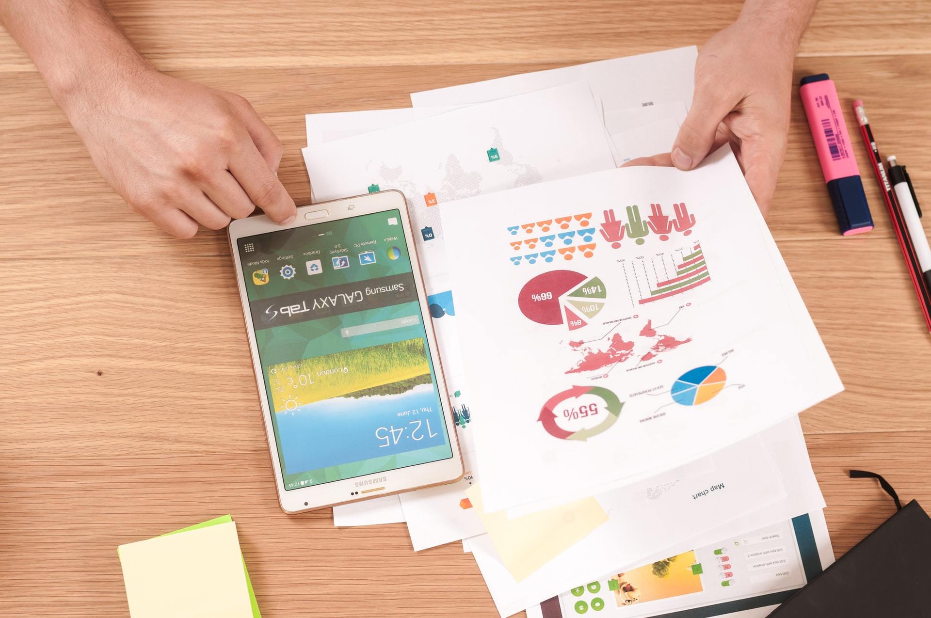 A person holds a sheet with colourful charts and graphs next to a tablet on a wooden desk with pens, highlighters, papers, and sticky notes scattered about.