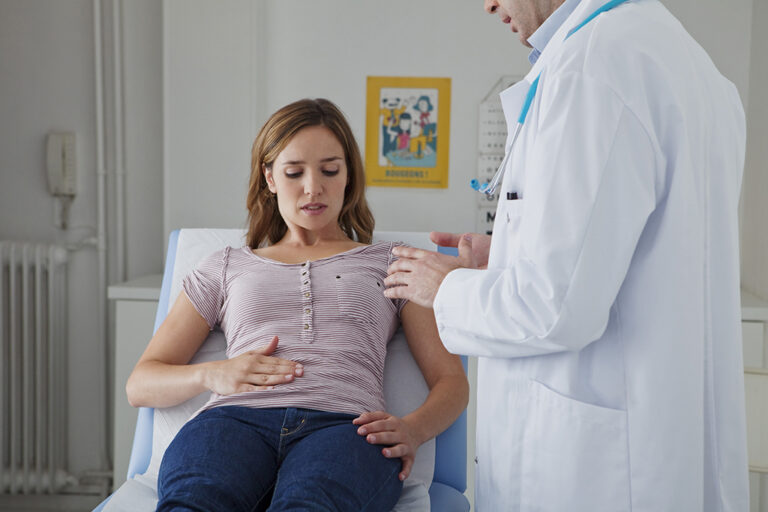 A doctor examines a woman sitting on an examination table. The woman looks concerned and touches her stomach while the doctor gently presses on her shoulder. They are in a surgery.