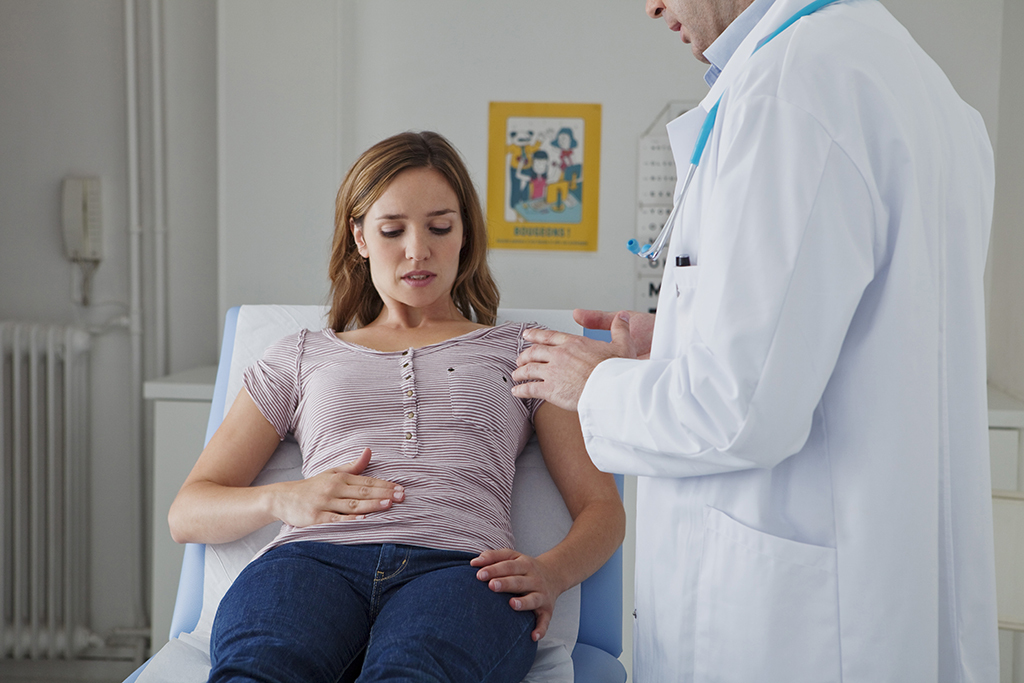 A doctor examines a woman sitting on an examination table. The woman looks concerned and touches her stomach while the doctor gently presses on her shoulder. They are in a surgery.