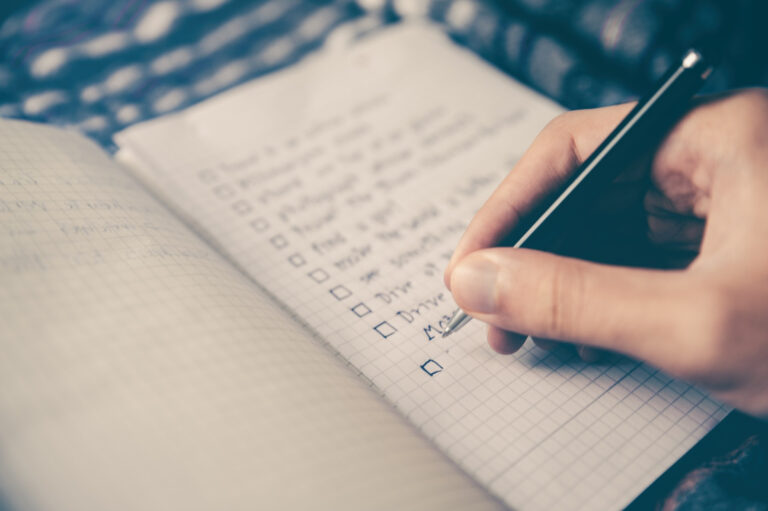 A hand holding a pen writes on a squared notebook with a checklist. Several items are already written, each next to an empty tick box. The background is slightly blurred.