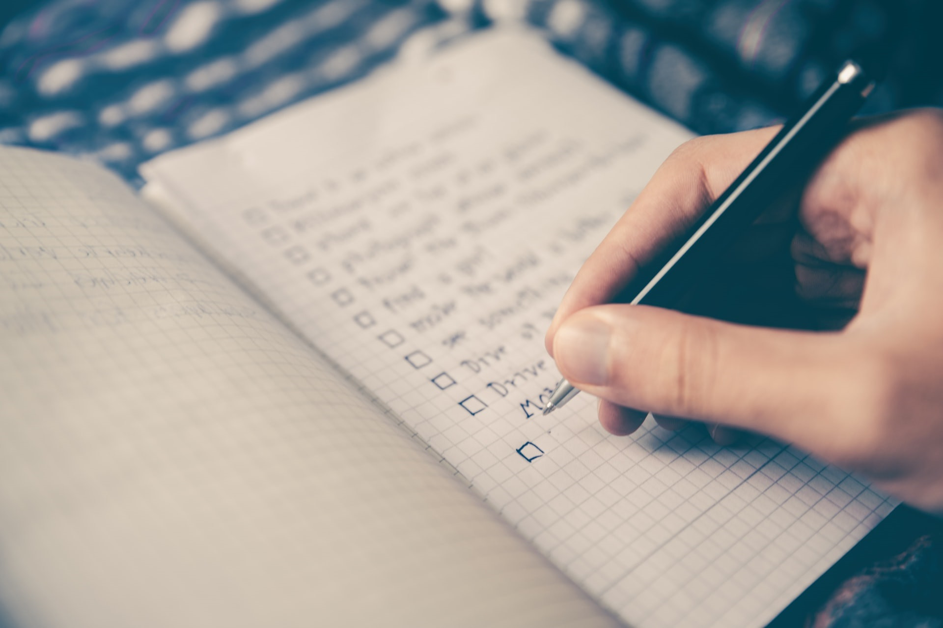 A hand holding a pen writes on a squared notebook with a checklist. Several items are already written, each next to an empty tick box. The background is slightly blurred.