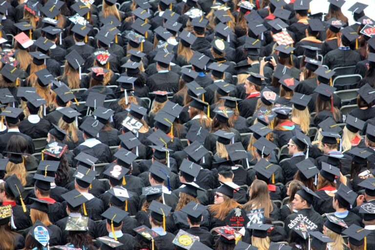 A large crowd of graduates wearing black caps and gowns sits outdoors. Many caps are decorated with colourful designs and messages. The image is taken from above, showing mostly the backs of the graduates.