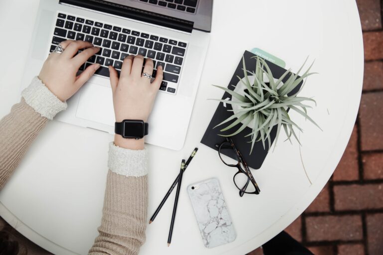 Person typing on a laptop at a round white table with a notebook, plant, spectacles, two pencils, and a marble phone case nearby. Only hands and part of arms are visible; workspace appears organised and modern.