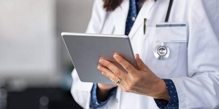 A doctor in a white coat with a stethoscope around their neck is holding and using a digital tablet.