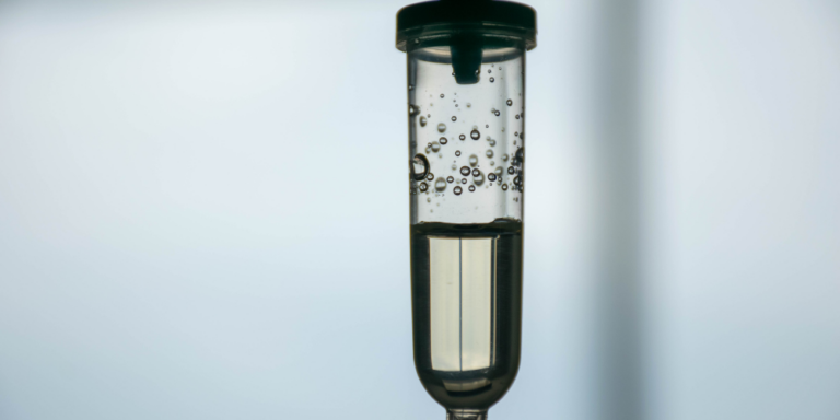 A close-up of a drip chamber filled with clear liquid and visible air bubbles, set against a blurred, light background.