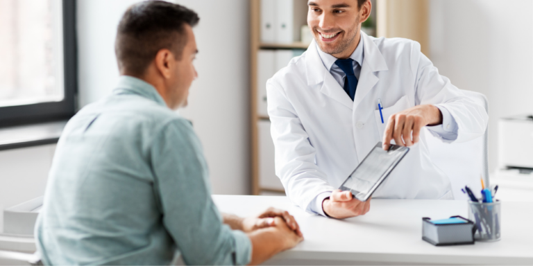 A doctor in a white coat shows a tablet to a patient sitting across the desk, both smiling and engaged in conversation in a bright office.