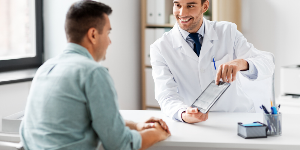 A doctor in a white coat shows a tablet to a patient sitting across the desk, both smiling and engaged in conversation in a bright office.