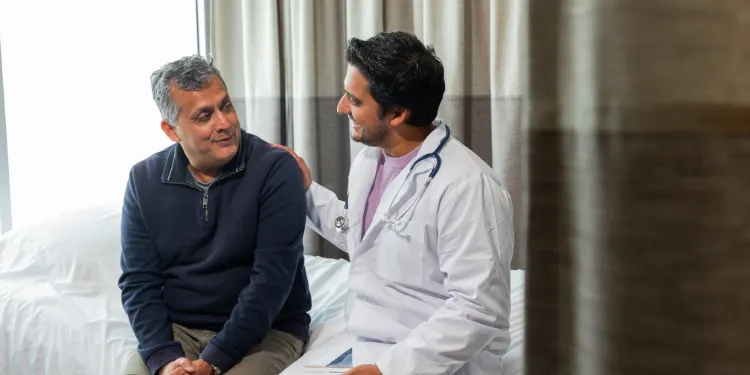 A doctor with a stethoscope comforts a seated patient, gently touching his shoulder as they talk in a bright medical surgery.