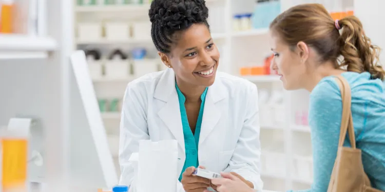 A pharmacist in a white coat smiles whilst assisting a customer at the counter. The customer holds a medicine box, and shelves with pharmacy items are visible in the background.