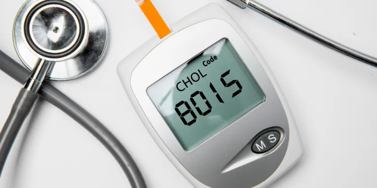 A cholesterol metre displaying a reading of 801.5, with a test strip inserted. A stethoscope lies next to the device on a white background.