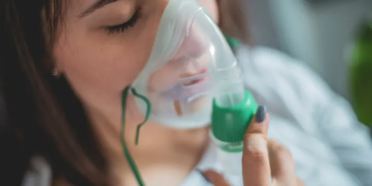 A person with closed eyes uses an oxygen mask, holding it gently to their face.