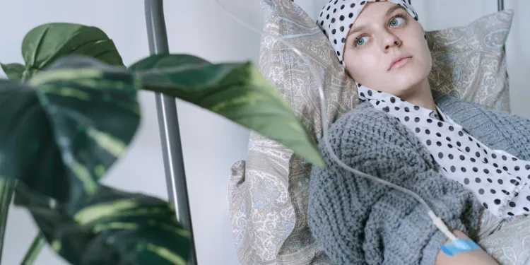 A person wearing a spotted headscarf lies in bed, receiving a drip, looking thoughtful. A leafy green plant is visible in the foreground.