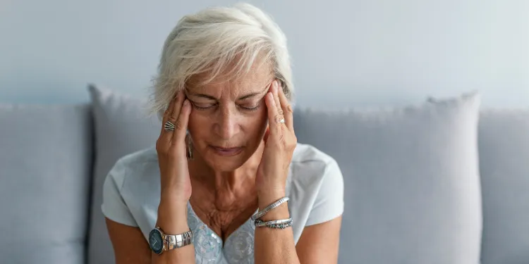 Older woman with short white hair sits on a sofa, eyes closed, holding her temples with both hands, appearing to have a headache or be in discomfort.