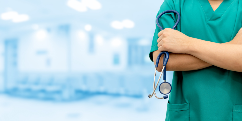A healthcare professional in green scrubs stands with arms folded, holding a stethoscope, in a blurred hospital or clinic setting.