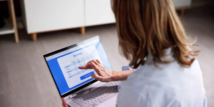 A person sits on the floor using a laptop, filling out an online survey form with a blue and white interface. Their finger is touching the screen, selecting an answer option.