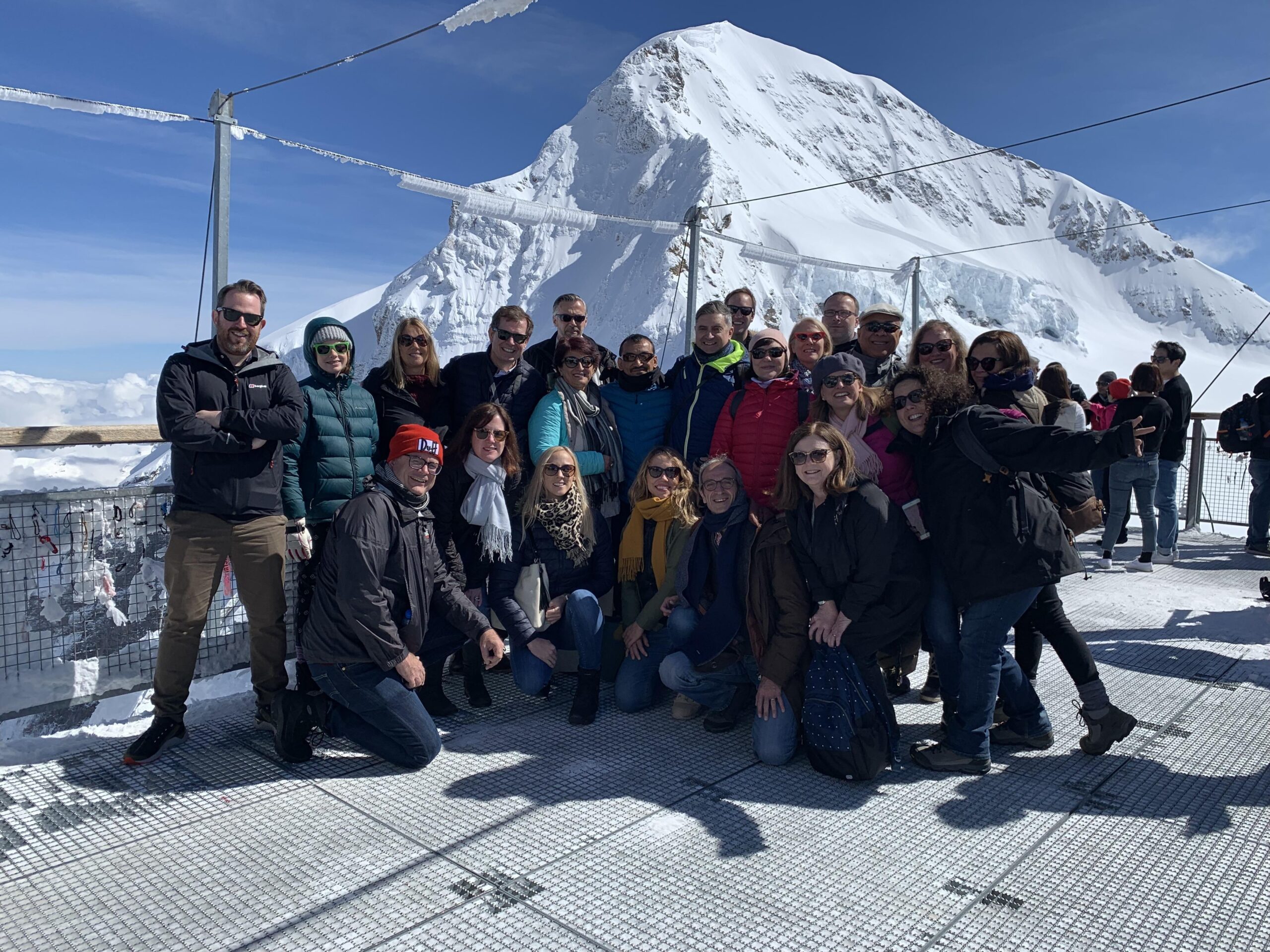 A group of people pose for a photo on a snowy viewing platform, with a large snow-covered mountain and blue sky in the background.