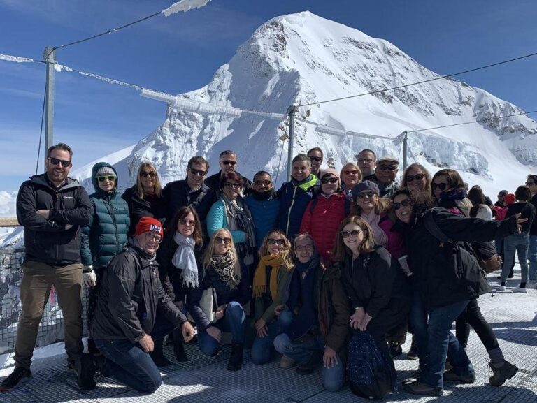 A group of people in winter clothing pose together on a snowy mountain platform with a snow-covered peak and blue sky in the background.