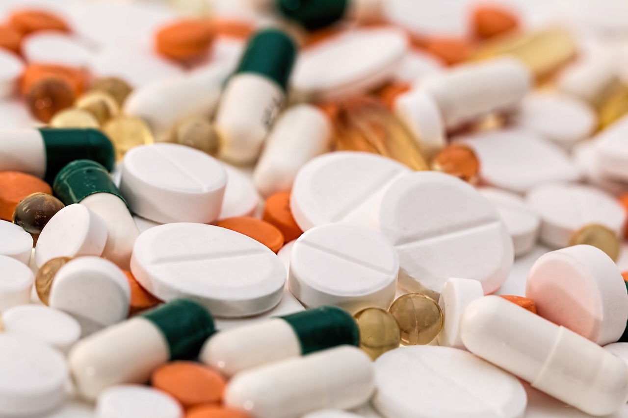 A close-up of various pills and capsules, including white tablets, orange tablets, green-and-white capsules, and yellow gel capsules, scattered in a pile.