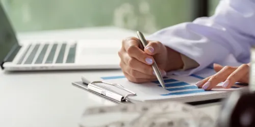 A person writes on a clipboard with a pen, analysing a blue bar graph. A laptop is open on the desk nearby.