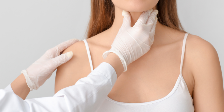 A doctor wearing gloves examines a woman's neck and throat area while she wears a white vest. The background is plain and light-coloured.
