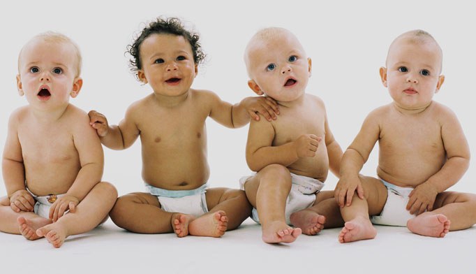 Four babies wearing nappies sit in a row on a white background, looking forward. They have different skin tones and expressions, and one baby has a hand on the shoulder of the baby next to them.