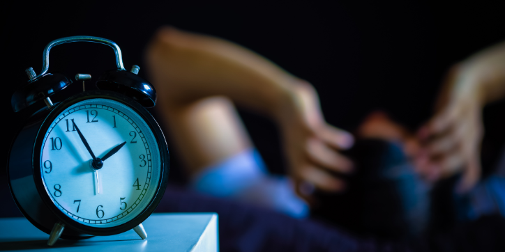 A classic black alarm clock shows 4.00 on a bedside table, with a blurred person in the background lying in bed, holding their head in apparent frustration or distress.