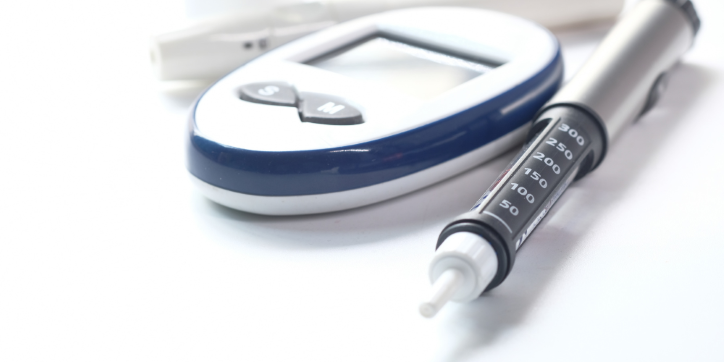 A glucose metre, a test strip, and an insulin pen are placed on a white surface, representing diabetes management tools.