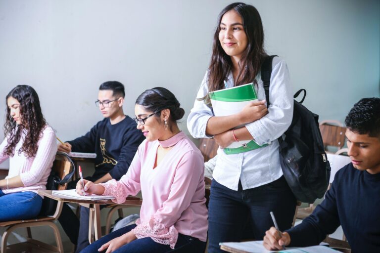 A student with a rucksack and book smiles while standing in a classroom as other students sit at desks, writing and studying.