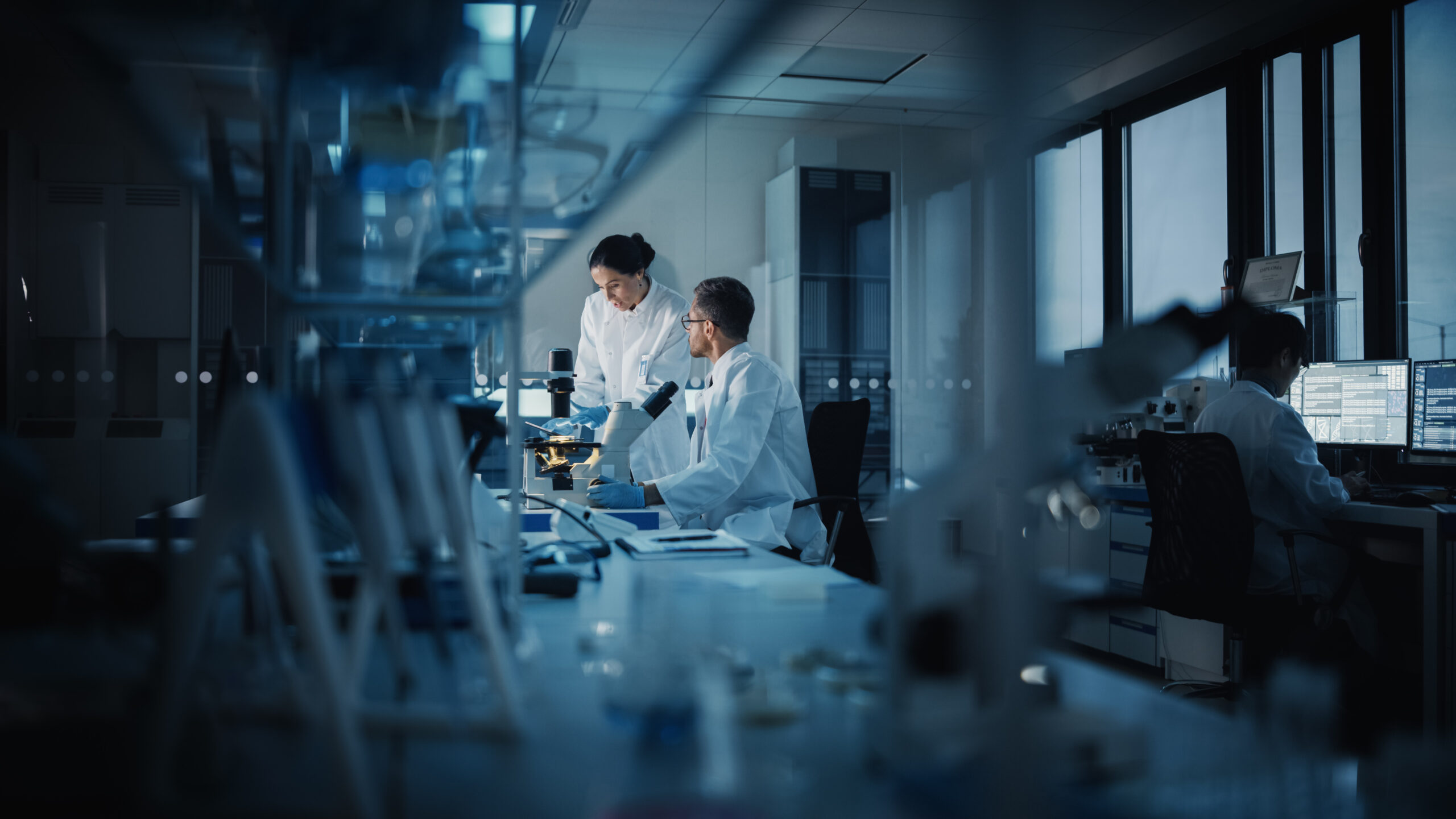 Two scientists in lab coats work together at a microscope in a modern, well-lit laboratory, whilst another person uses a computer in the background. Scientific equipment and glassware are visible throughout the room.