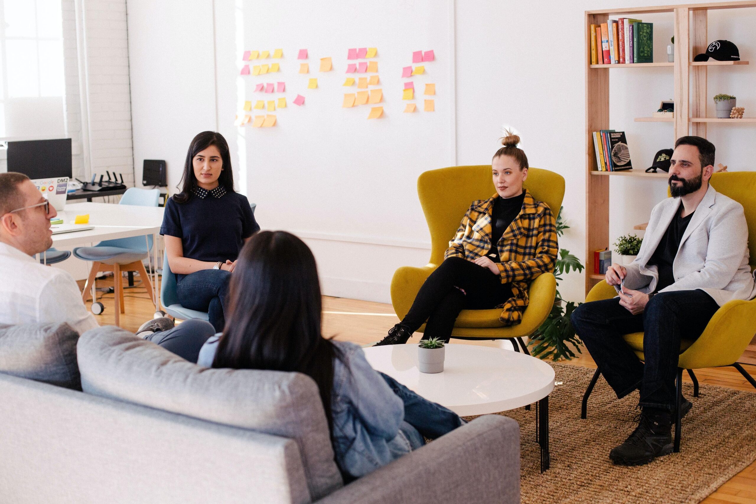 Five people sit in a circle in a modern office, engaged in discussion. There are colourful sticky notes on the wall and bookshelves in the background. The atmosphere appears collaborative and casual.